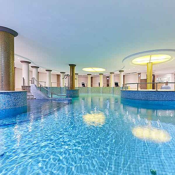 Das Bild zeigt einen großen, hellen Innenpool, der von Säulen und Mosaikfliesen umgeben ist. Das Wasser des Pools ist klar und hellblau, und die Oberfläche ist ruhig und spiegelglatt. Im Hintergrund befinden sich mehrere Säulen, die einen Eindruck von Größe und Eleganz vermitteln. An den Rändern des Pools sind einige Liegen aufgestellt, die auf Entspannung und Erholung hindeuten. Eine kleine Rutsche ist ebenfalls in der Nähe des Pools zu sehen, was darauf hindeutet, dass der Pool sowohl für Erwachsene als auch für Kinder geeignet ist. Die allgemeine Atmosphäre des Bildes ist ruhig, friedlich und entspannend. Das Licht ist weich und diffuses, was zu einem beruhigenden und einladenden Ambiente beiträgt. Die Farben sind sanft und beruhigend, hauptsächlich Blau- und Goldtöne, die auf Luxus und Ruhe schließen lassen. Die Gesamtstimmung des Bildes ist eine von Frieden, Gelassenheit und Wohlbefinden.