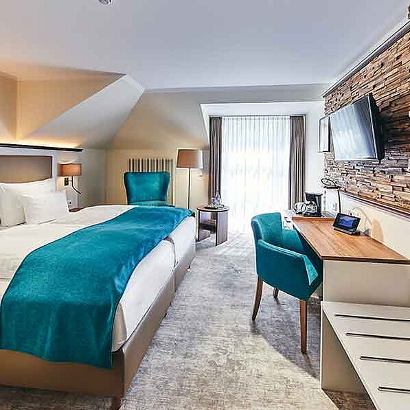 Das Bild zeigt ein geräumiges und stilvoll eingerichtetes Hotelzimmer. Im Mittelpunkt steht ein großes Doppelbett mit türkisfarbener Tagesdecke. Das Bett ist mit weißen Bettwäsche und Kissen bezogen. Neben dem Bett steht ein kleiner Nachttisch mit einer Tischlampe. Auf der gegenüberliegenden Seite des Raumes befindet sich ein Schreibtisch mit einem modernen Design aus hellem Holz. Ein bequemer Sessel in der gleichen türkisfarbenen Farbe wie die Tagesdecke steht vor dem Schreibtisch. An der Wand hinter dem Schreibtisch ist ein moderner Flachbildschirmfernseher angebracht. Die Wand selbst ist mit einer Holzverkleidung in warmen Brauntönen gestaltet, die ein natürliches und gemütliches Ambiente schafft. Ein weiterer Sessel in der gleichen Farbe steht weiter im Raum, und ein kleiner Beistelltisch steht zwischen den beiden Sesseln. Ein großes Fenster mit weißen Vorhängen lässt viel Tageslicht in das Zimmer und bietet einen Blick auf ein begrüntes Umfeld (obwohl die Details nicht klar zu erkennen sind). Das Zimmer ist hell und freundlich eingerichtet, die Atmosphäre wirkt ruhig und entspannend, ideal für einen komfortablen Aufenthalt. Die Farben sind harmonisch aufeinander abgestimmt, in sanften Naturtönen und erfrischendem Türkis. Insgesamt vermittelt das Bild einen Eindruck von modernem Komfort und stilvoller Eleganz.
