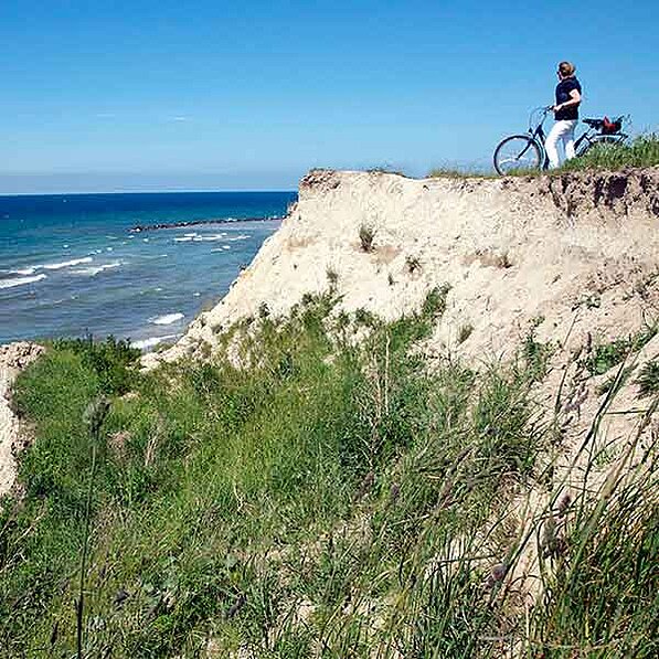 Das Bild zeigt eine Frau, die auf einer sandigen Klippe steht und auf das Meer blickt. Sie trägt eine dunkle Oberbekleidung, eine helle Hose und scheint bequemes Schuhwerk zu tragen – typische Kleidung für eine Radtour. Ihr Fahrrad steht neben ihr, auf dem Boden der Klippe. Das Fahrrad sieht aus wie ein Tourenrad oder ein Hybridrad, robust genug für unebenes Gelände, aber komfortabel für längere Fahrten.
Der Vordergrund besteht aus kurzem, grasigem Bewuchs, der die Klippe bedeckt. Der Hintergrund zeigt eine klare blaue See oder Ozean mit sanften Wellen. Es ist ein sonniger Tag, der Himmel ist fast wolkenlos und hellblau. Die allgemeine Atmosphäre ist entspannt und ruhig; es herrscht eine friedliche Stimmung. Das Bild strahlt eine gewisse Ruhe und die Freude an der Natur aus. Die Klippe selbst erscheint sandig und etwas erodiert, was auf natürliche Abtragungsprozesse hindeutet. Die Farben des Bildes sind natürlich und leuchtend, mit dem Blau des Meeres als dominantem Farbton, kontrastiert durch das Grün der Vegetation und das Sandgelb der Klippe. Das Bild vermittelt ein Gefühl von Freiheit und einem idealen Ort zum Entspannen und die Aussicht zu genießen.